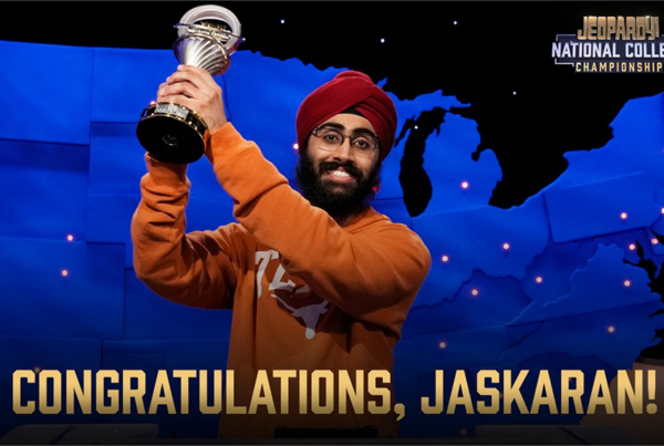 a man wearing a red turban and orange University of Texas sweatshirt holding a trophy he won at Jeopardy, with a blue map of the united states in the background of the jeopardy set he's standing on