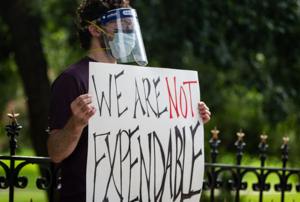 a man wearing a face shield and face mask standing in front of a wrought iron fence with a sign that says we are not expendable--a protest by teachers against state education rules during the pandemic
