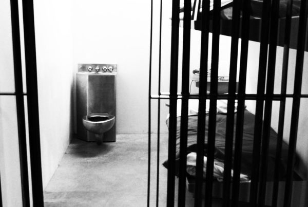 a black and white photo of a prison cell with a bed and a metal toilet in the corner