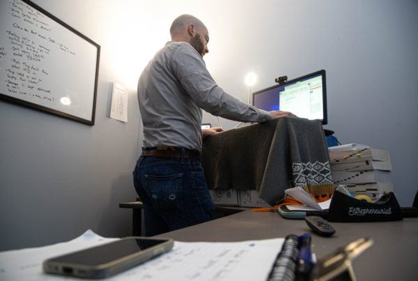 a man in a button down shirt and slacks stands at a desk looking at his computer screen where he is having a video conference session