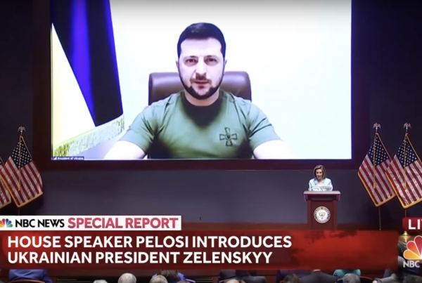 a man wearing an army green t shirt sitting in a brown office chair with the ukrainian flag behind him, appearing on a large video screen addressing a large auditorium with members of the U.S. congress