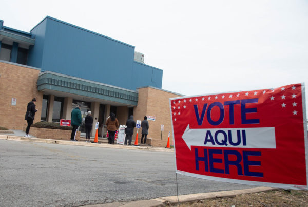 In the foreground, a large sign indicating the polling location. In the distance is a line of people outside a building.