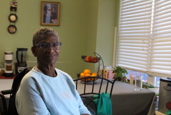 a Black woman with short-cropped hair, stylish blue glasses and a white sweatshirt sitting at a chair near her kitchen table