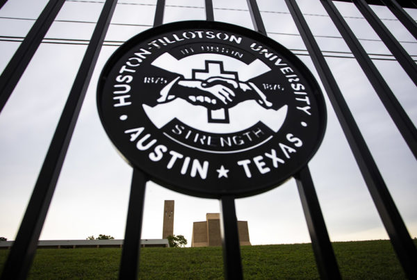 an iron fence with a seal in it with two hands shaking and a cross behind them, with the words huston tillotson university austin texas around the seal and the years 1875 and 76