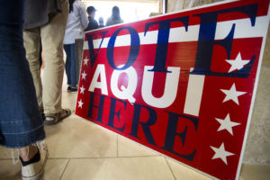People stand in line next to a red sign that says "vote here/aqui"