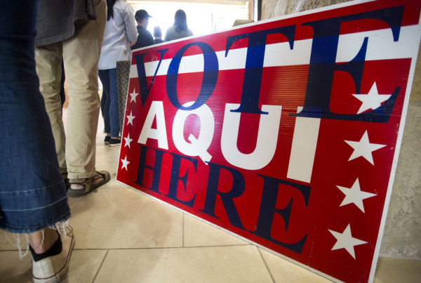 People stand in line next to a red sign that says "vote here/aqui"