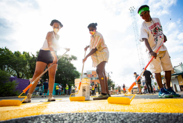 A low angle photo shows three people using long rolling brushes to paint yellow lettering on a street.