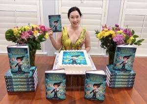 A photo of a woman sitting behind a large table stacked neatly with hardcover copies of the book "Lia Park and the Missing Jewel." The author holds one book and smiles at the camera. Also on the table are colorful flower bouquets and a cake with the book cover image printed on it.