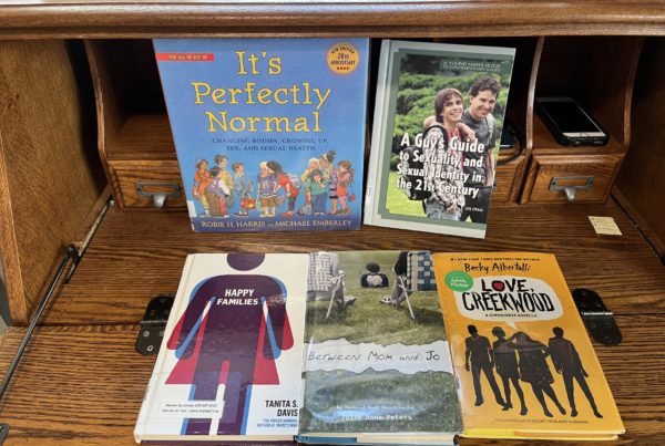 Books on a table include "It's Perfectly Normal," "A Guy's Guide to Sexuality and Sexual Identity in the 21st Century" and "Between Mom and Jo"
