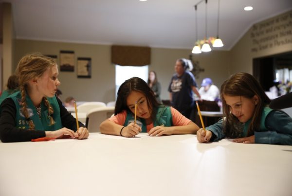 Three girls wearing green Girl Scout vests sit at a table, each writing on a sheet of paper
