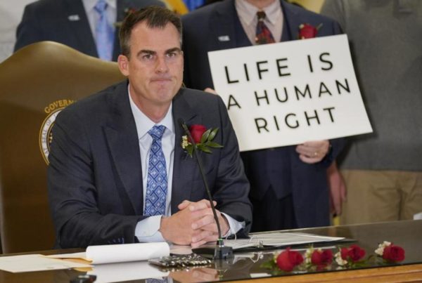 Oklahoma governor Kevin Stitt sitting at a desk while someone behind him holds a sign that says "life is a human right"