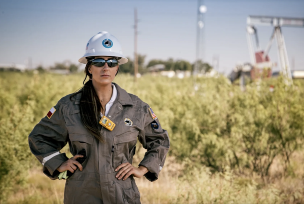 Sarah Stogner stands outside with her hands on her hips, wearing a hard hat, sunglasses and jumpsuit. A pump jack is in the background.