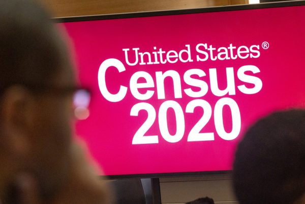 A screen displays "United States Census 2020" in white letters on a red background. People stand in the foreground in front of the screen.