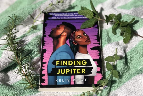 A book sits on a green and white striped beach towel, surrounded by trimmings of herbs. The book cover features two black teenagers leaning with the backs against one another.