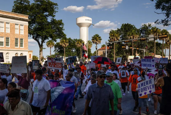 People solemnly march through a street carrying signs and photos. A water tower reading Uvalde is in the background.