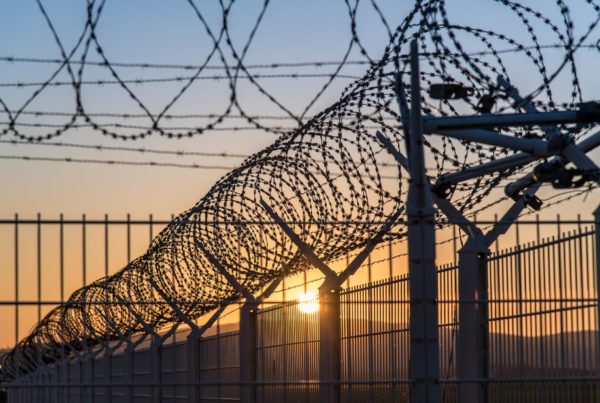 A few rows of razor wire and fencing are shown against a setting sun.