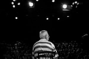 A black and white photo of a man from behind. The man is wearing a striped shirt and sitting in a chair on a stage facing a large audience and studio lights.