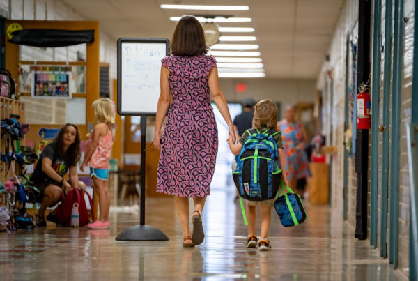 An adult holds the hand of a child wearing a backpack and carrying a lunchbox through the hall of an elementary school.
