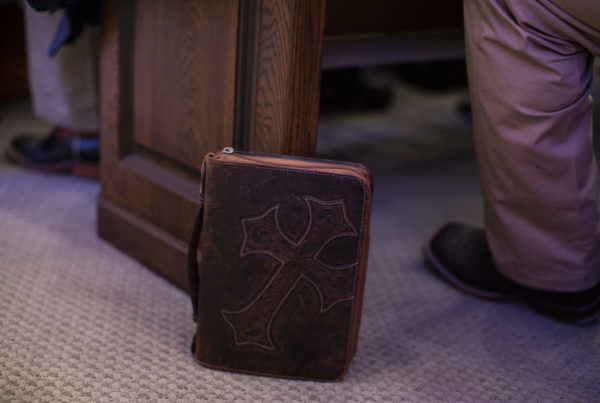 A bible in a leather case with a cross on it rests against a church pew next the foot of a person wearing cowboy boots.