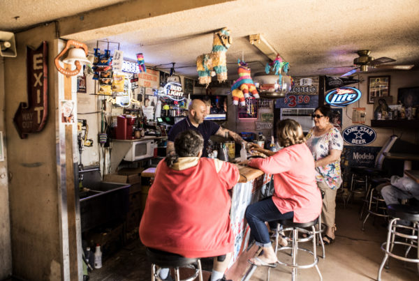 A photo of the inside of a small bar with piñatas hanging from the ceiling and memorabilia covering the walls. Three people are gathered around the counter as a bartender helps them.
