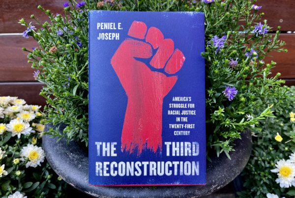 A hardcover copy of the book "The Third Reconstruction" rests against a backdrop of potted flowers. The cover of the book is dark blue with a red fist image on it.