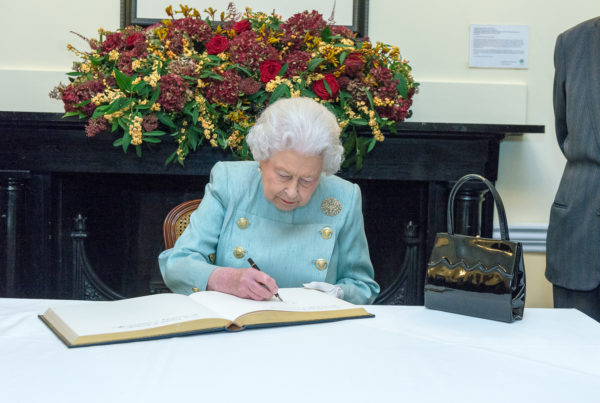 The queen sits at a table, outdoors, writing in a book