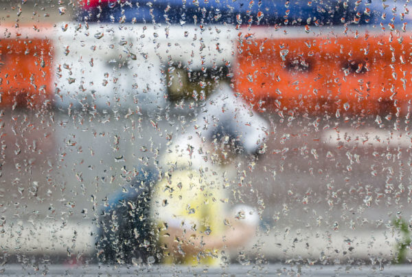 A photo shot from behind a rain-streaked window shows a person walking through wet weather wearing a clear poncho