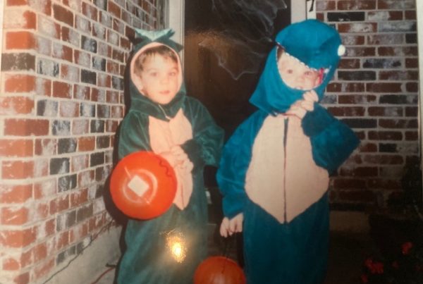 Two little kids wearing homemade Halloween costumes hold plastic pumpkins in front of a brick home.