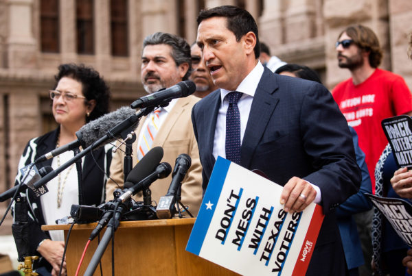 Rep. Trey Martinez Fischer, in a navy blue suit, speaking at a podium outside the Texas Capitol. He's holding a signthat reads "Don't mess With Texas Voters."