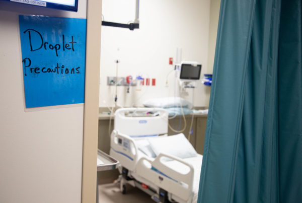 A photo peeking past a green curtain into an empty hospital room. On the wall by the room is a sign that says "droplet precautions."