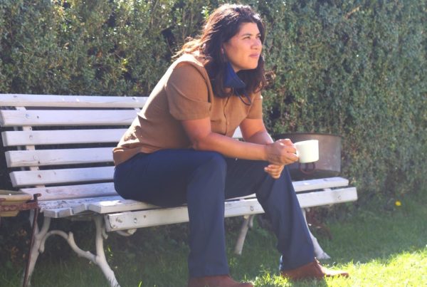 mónica teresa ortiz sits on a white bench holding a white coffee cup. There's greenery in the background.