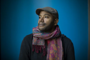 A photo of Peniel Joseph. He wears a colorful scarf over a black shirt and a hat. He looks to the left with a serious face. The background is blue.