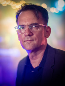 A close-up photo of Thom Zimny. He looks at the camera. He wears a black shirt and glasses.