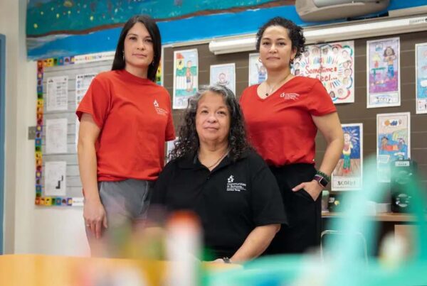 A portrait of three women in a classroom: One sitting, wearing a black shirt, and two wearing red shirts stand on either side of her. Their shirts say "Communities in Schools"