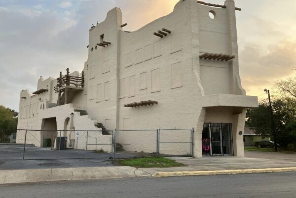 A long, tan building is seen. It's viewed from across the street and off to the side so the entrance and the side of the building is seen. This is the Aztec Building, where the Texas Conjunto Music Hall of Fame and Museum is now housed.