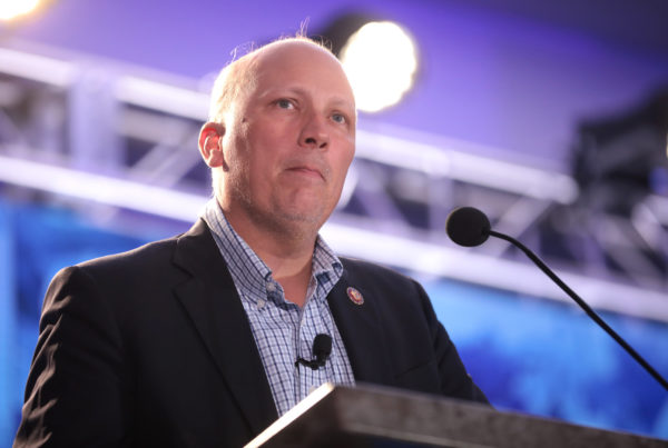 Texas Republican Congressman Chip Roy standing at a lectern with a microphone near his mouth, wearing a dark blazer and checked button-up shirt.