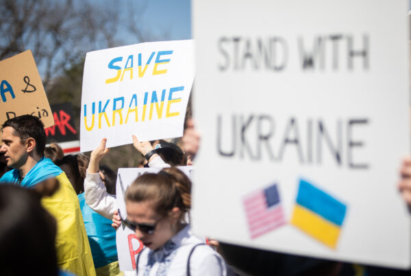 Demonstrators gather, holding protest signs. Two prominent signs in the image read "Save Ukraine" and is colored in the yellow and blue of the Ukrainian flag. In the foreground another sign reads "Stand With Ukraine" and shows the U.S. and Ukrainian flags crossed together beneath the words.