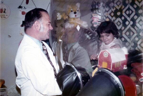 A 1970s photo of a young dark-haired boy in a plastic enclosure playing with a toy barn. A doctor interacts with him outside of the plastic.