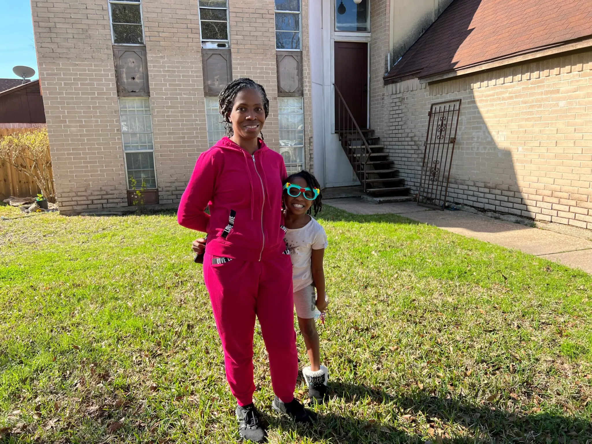 A woman stands on a lawn in which a brick building is seen the background. She's smiling and next to her is a young girl wearing teal-framed sunglasses. Both are smiling for the camera. This is Kimberly Lee and her granddaughter.