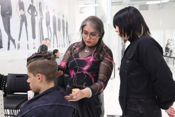 Three people are seen in a salon setting. A client is seated to the right of the photo wearing a chair cloth and facing to the right with his hair held up with clips. Next to him an instructor holding clippers gestures at his hair in mid-conversation with a student who looks on.