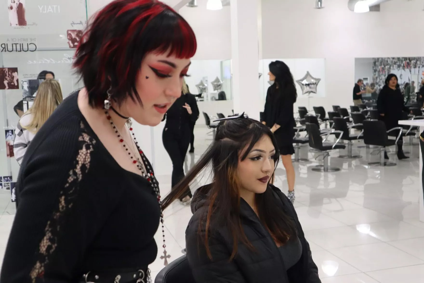 Two people are seen in a salon setting. One person is seated and covered with a chair cloth while a cosmetology student prepares tinsel for her hair in the foreground.