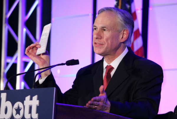 Governor Greg Abbott is seen speaking at a podium, holding up a pamphlet. His name is fixated on the podium, suggesting this is a campaign event.
