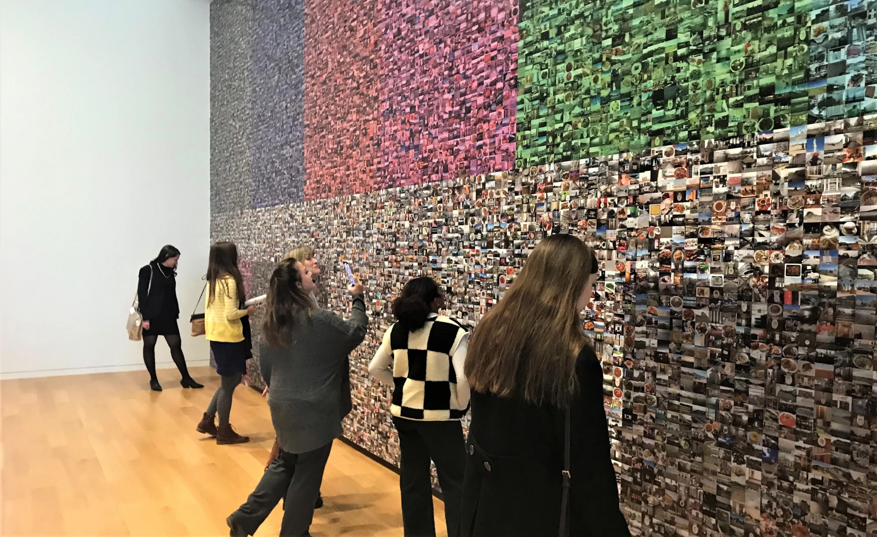 Five museum-goers look at and take cell phone photos of the thousands of small images that make of Hasan Elahi's 
