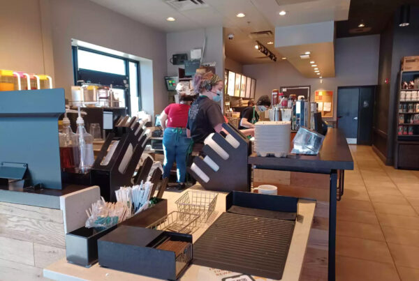 The interior of a Starbucks is seen. One worker stands behind a register wearing a face mask and a second worker is seen from behind at another register by the drive-thru window. A third worker wearing a beanie and face mask is also seen in the distance.