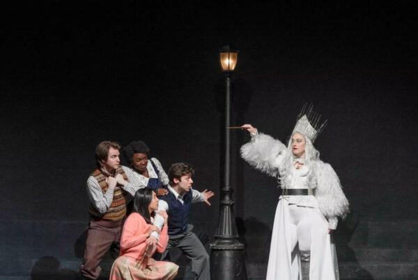 Actors on stage perform a scene from a production of The Lion, the Witch, and the Wardrobe. Four of the actors stand on the left, crouching and backing away from the actor dressed as the White Witch, who is clothed in a costume of all white, including a crown, and is holding up a wand pointed at the group as if about to cast a spell.