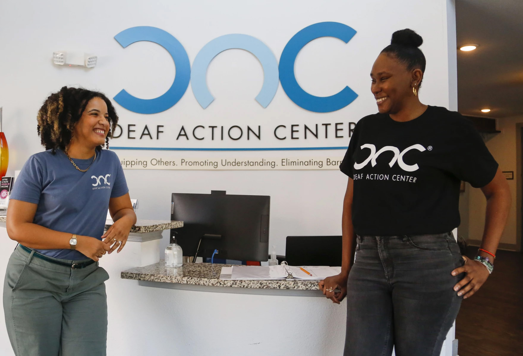 Two women stand on either side of the photo, facing each other and smiling. They're standing in front of what looks to be the reception desk of the Deaf Action Center, the logo of which is seen on the wall behind them as well as on their t-shirts. On the left is Laura Tovar, development director of the center, and on the right is Bianca Walker, a deaf and hard of hearing specialist at the center.