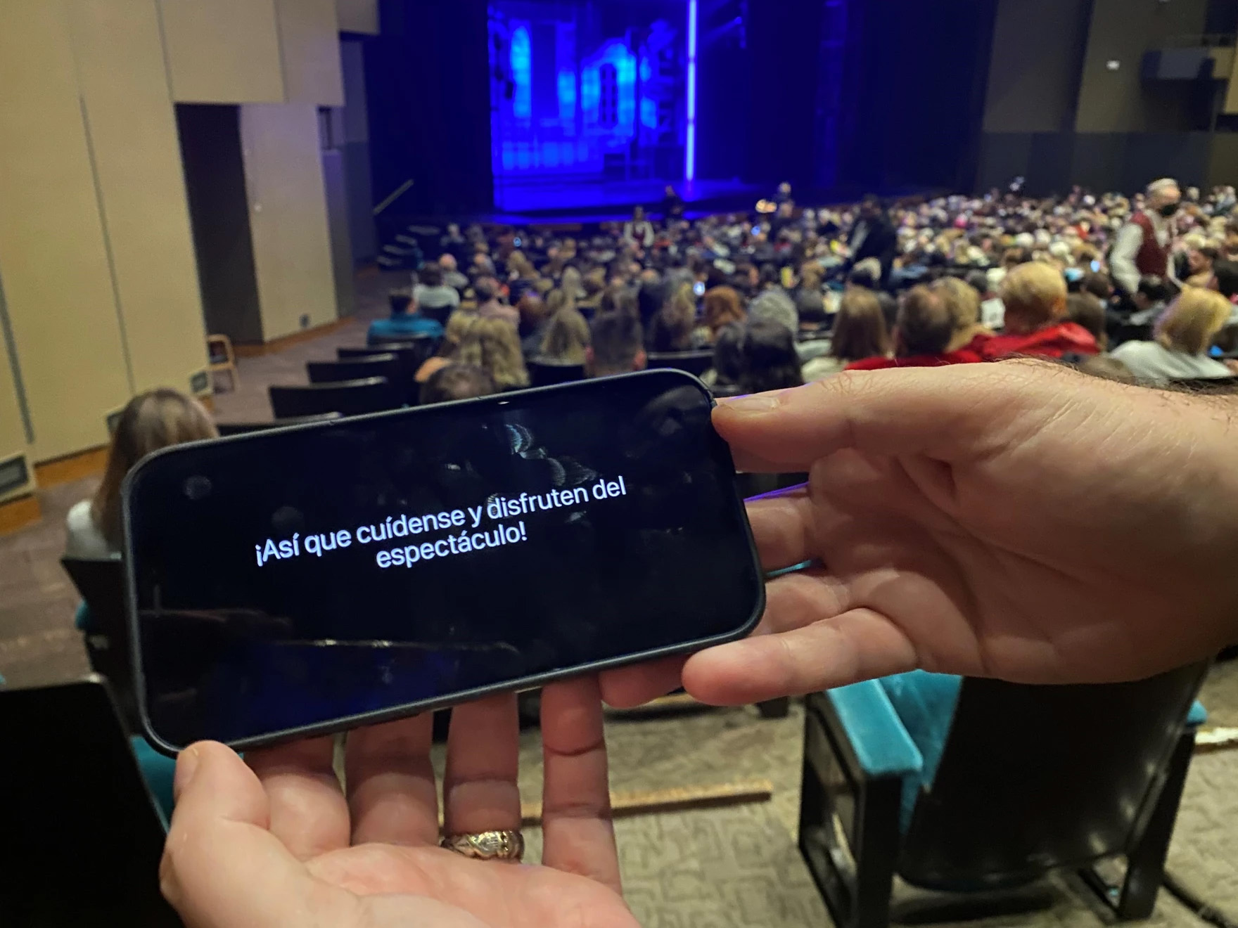A closeup of someone's smartphone showing the captions of the theater production they're watching. The stage can be seen in the distance behind the phone. The caption is in Spanish, reading 