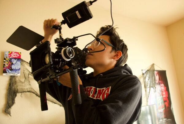 A young man handles filmmaking equipment. He's holding up the camera and gazing up at an external appendage. This is Malon Rubio Smith.