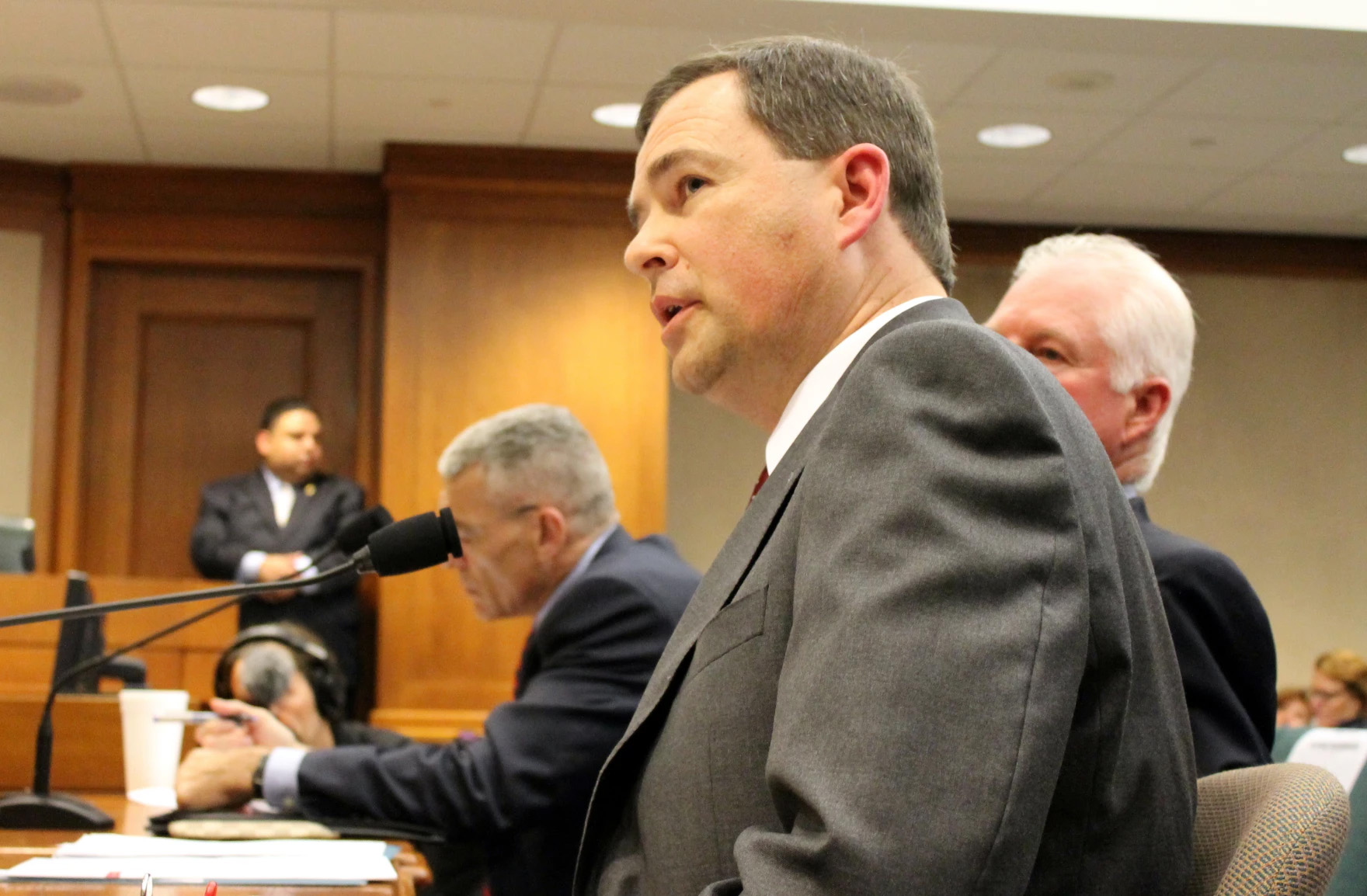 A seated man dressed in a suit is seen from the side speaking into a microphone. He appears to be in a courtroom. This is Brandon Woods, executive director of the Texas Commission on Jail Standards.