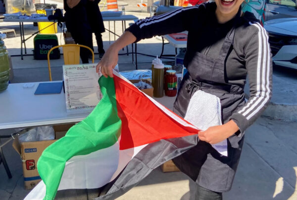 A woman faces the camera smiling as she unfurls a Palestinian flag. She's wearing an apron and tables with condiments are seen behind her. She's outside. This is Sara Masoud.